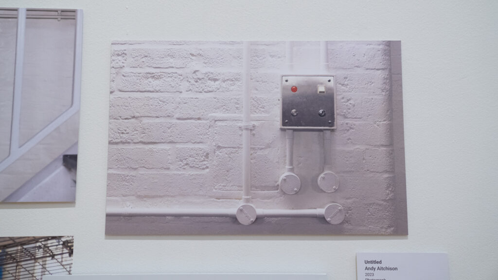 Outside a cell doorway at HMP/YOI Lincoln, the shape of a filled-in candle alcove is visible in the painted brickwork underneath the electric cabling for an alarm. 2022.